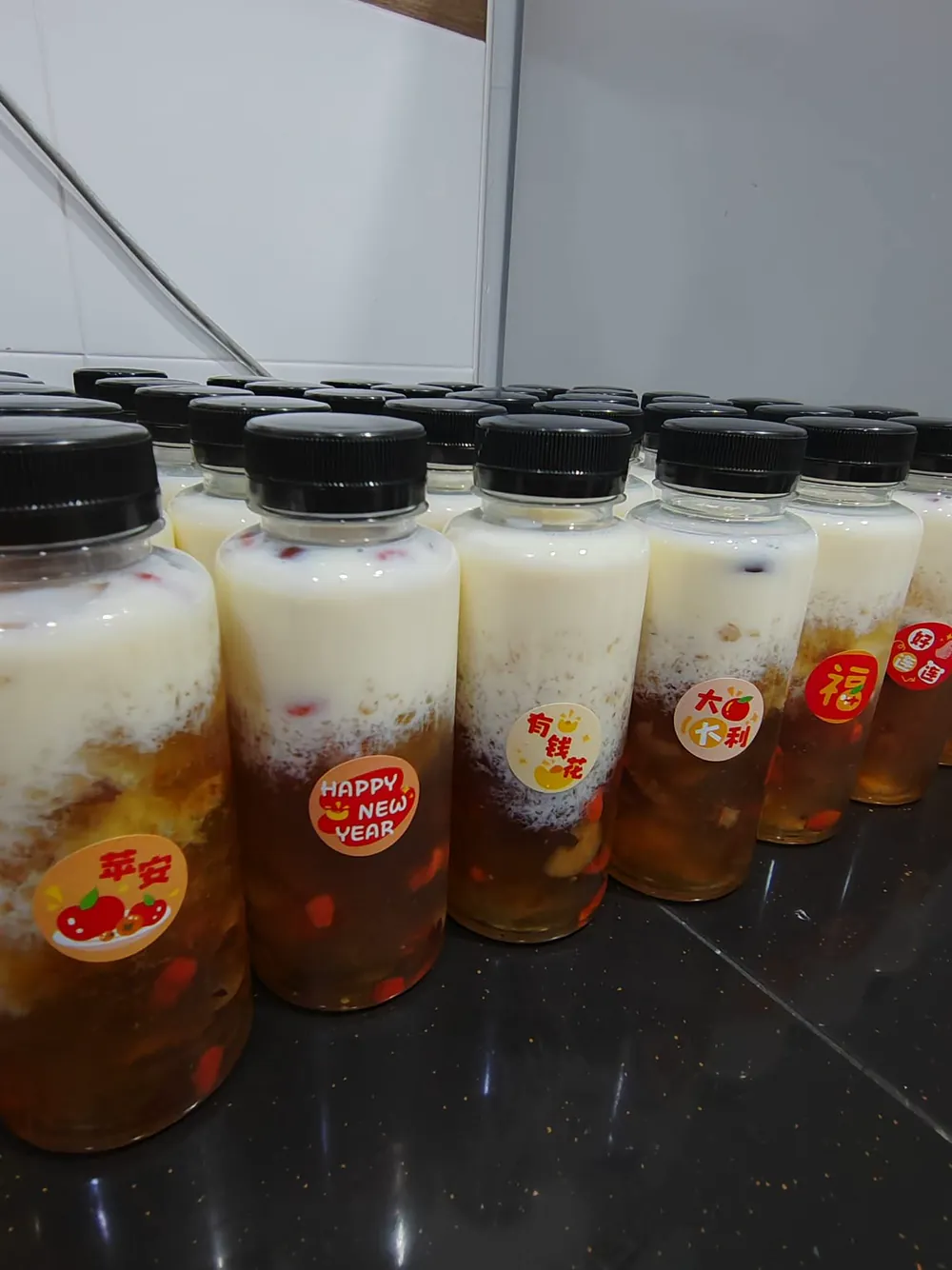 一排桃胶雪燕牛奶，瓶身贴着平安、有钱花、大吉大利、福等祝福贴纸。