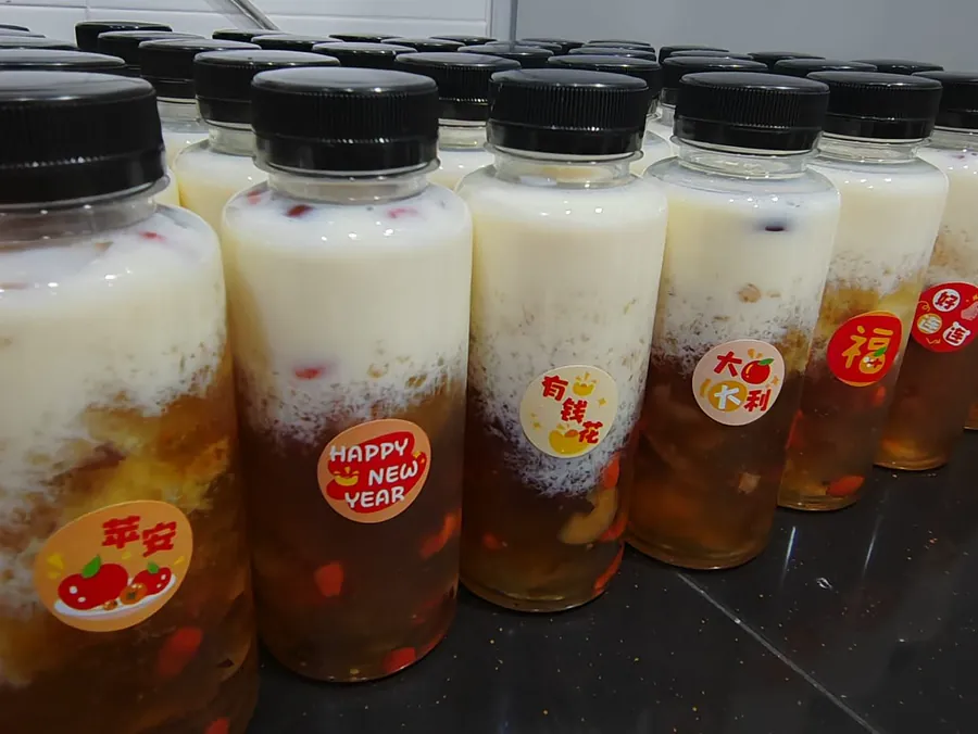 一排装好瓶的桃胶雪燕牛奶，瓶身贴着平安、Happy New Year、有钱花、大吉大利、福的贴纸。